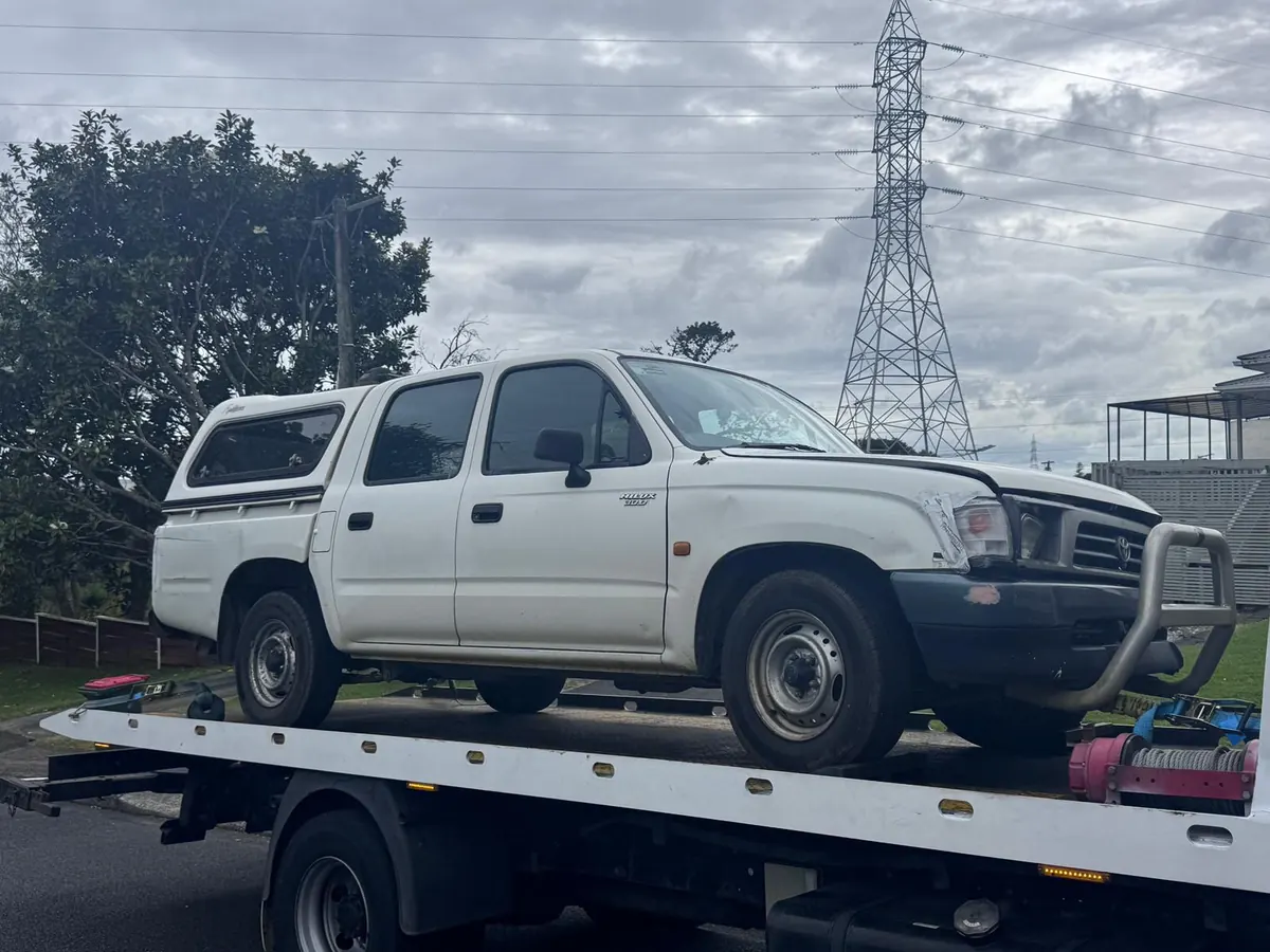 Damaged white Toyota Hilux being collected on a flatbed tow truck in South Auckland